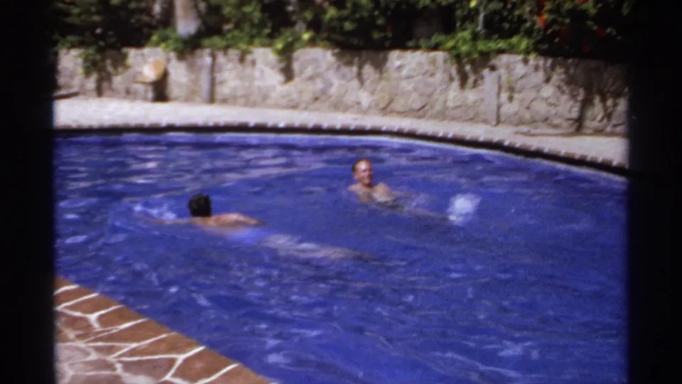 1974 Cabo San Lucas home-movie frame showing two swimmers in a bright blue outdoor pool bordered by stone walls and plants.