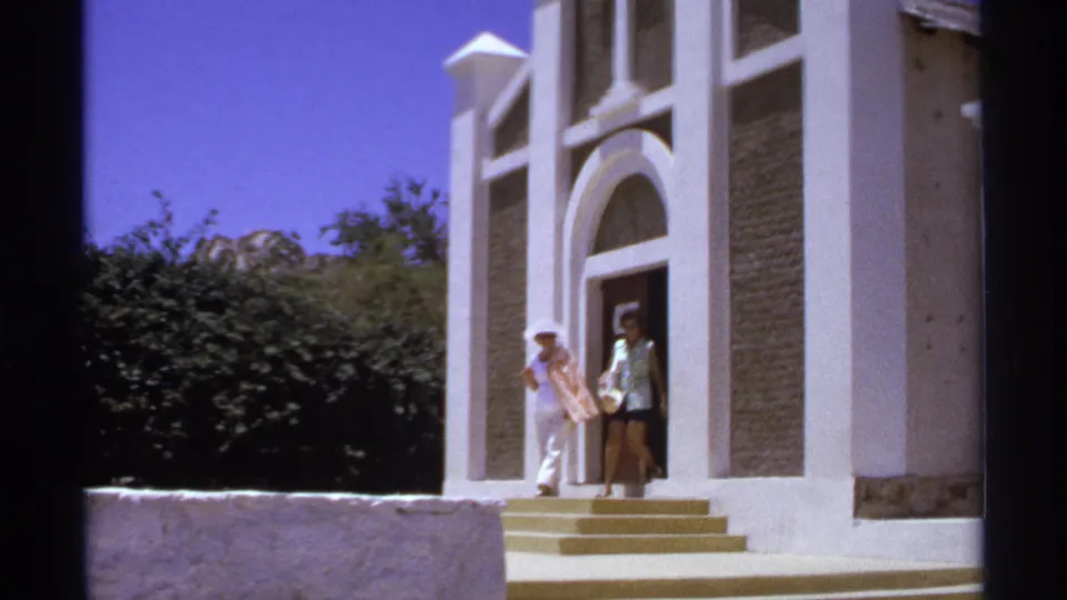 1974 Cabo San Lucas frame with two women in light clothing near a white church entrance with arched doorway and steps.