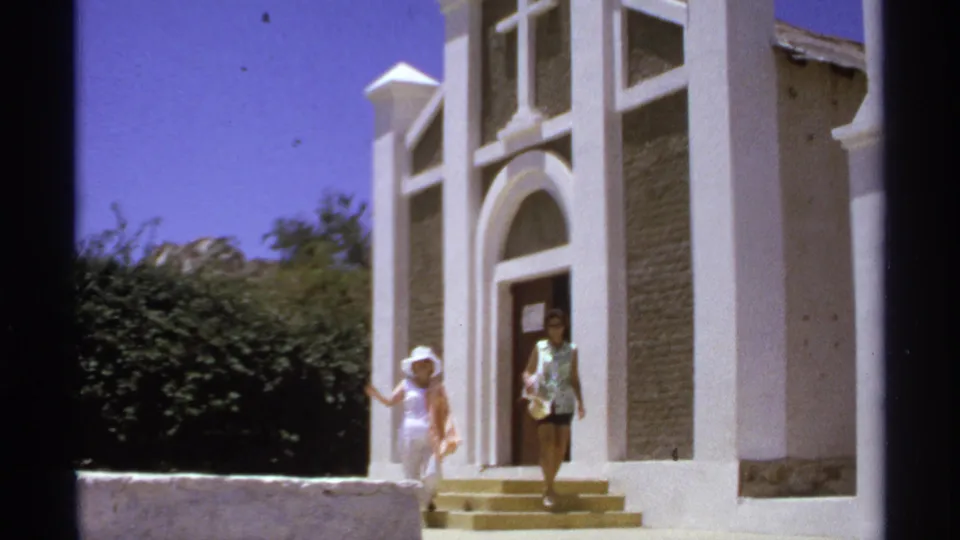 1974 Cabo San Lucas frame showing a woman leaving a white church exterior while another figure stands at the arched doorway.