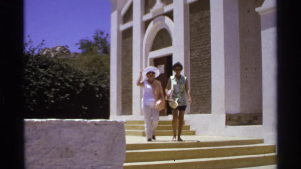 The women continue down the church steps in 1974 Cabo San Lucas, one wearing a white hat and the other a patterned top with dark shorts.