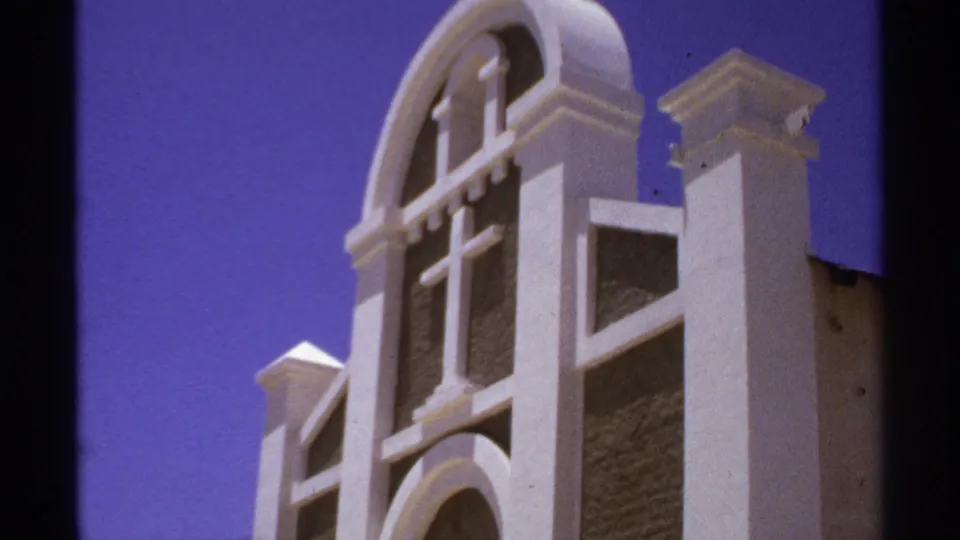 Close 1974 8mm view of a white church facade in Cabo San Lucas, showing the cross, arched details, and dark recessed panels.