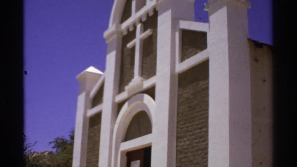 1974 Cabo San Lucas 8mm frame showing the church facade in sunlight, with white stucco pillars, arched trim, and a central cross.