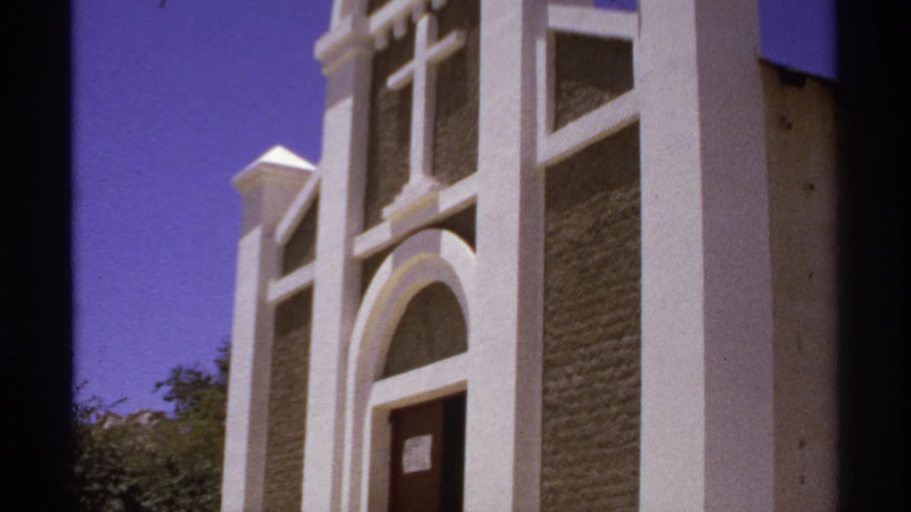 White church facade with arched doorway, cross, trees, and blue-purple sky.