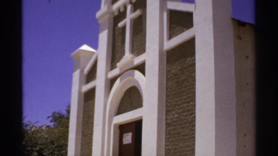 1974 Cabo San Lucas 8mm frame showing a white church exterior with arched doorway, cross, textured walls, trees, and saturated sky.