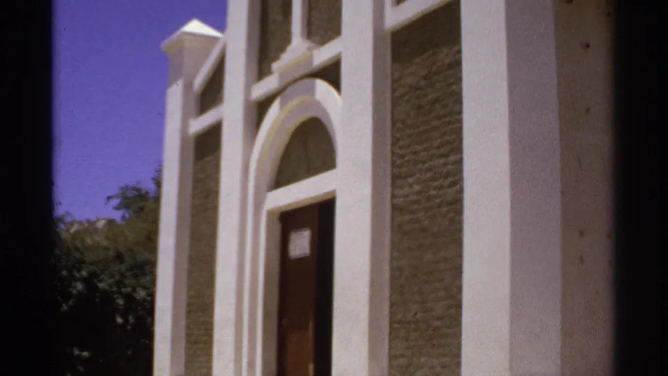 1974 Cabo San Lucas frame with the church entrance shifted in view, showing arched white trim, textured walls, door, cross, and foliage.