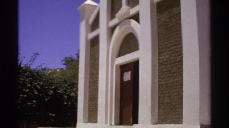 1974 Cabo San Lucas 8mm frame showing the lower church facade, arched doorway, dark door, textured wall panels, trees, and saturated sky.