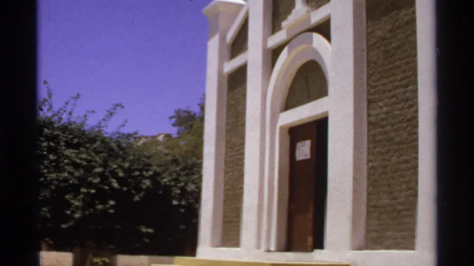 1974 Cabo San Lucas frame showing the white church facade from a visitor angle, with arched doorway, cross, textured wall panels, trees, and film edge.