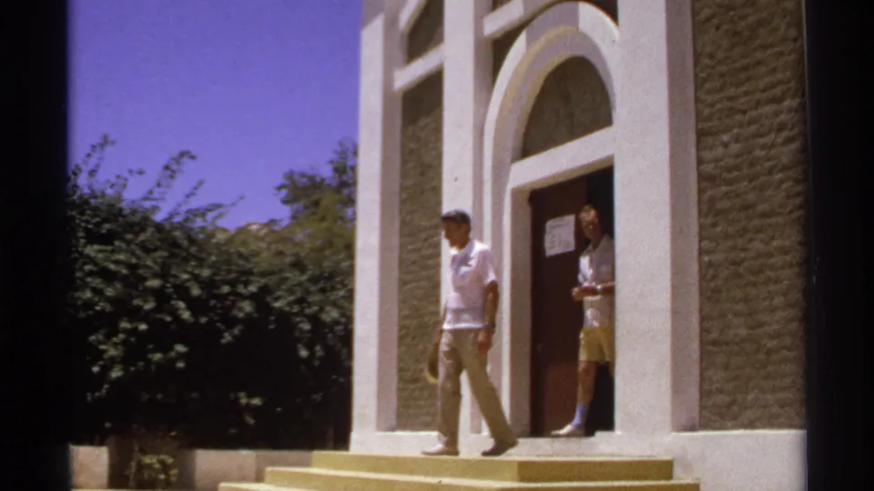 Two adult men in light summer attire are visible at the arched entrance, with one already walking down the steps in the 1974 Cabo San Lucas footage.