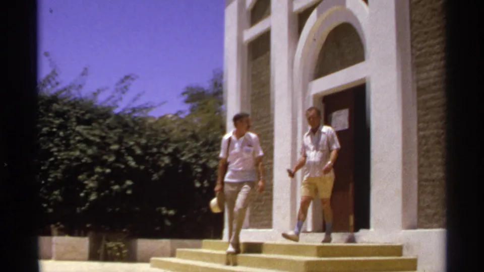 Two adult men descend the yellow steps outside a white arched building, one carrying a light-colored hat, in the supplied 1974 Cabo San Lucas scene.
