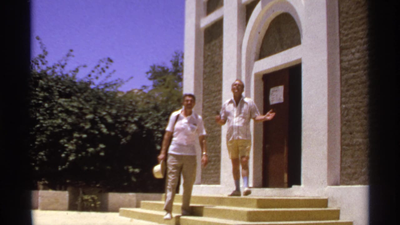 Two men stand outside a white stucco building with arched doorways and steps.