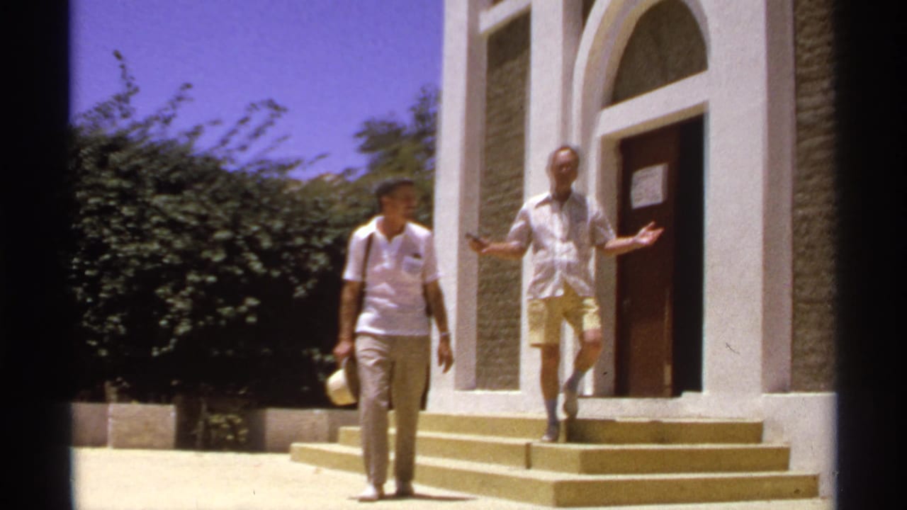 Two men walk down steps from a stucco building, one carrying a straw hat.