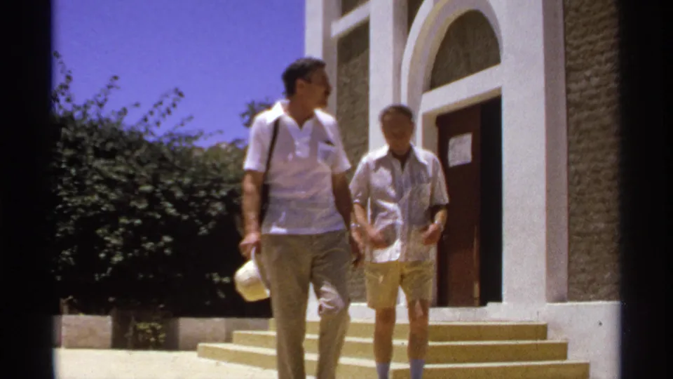 Two adult men pass in front of the white arched building in Cabo San Lucas, with the nearer man turned slightly to the side while walking.