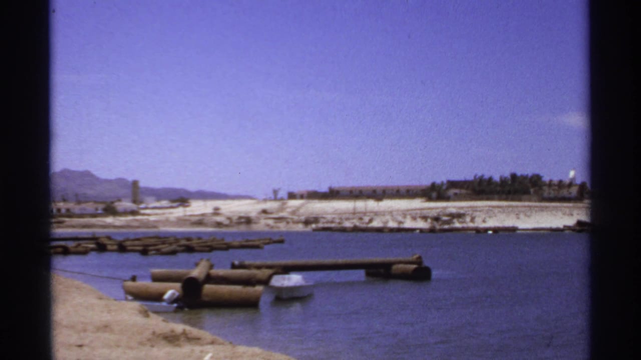 Calm water, log docks, sandy shoreline, and distant low buildings under blue sky.