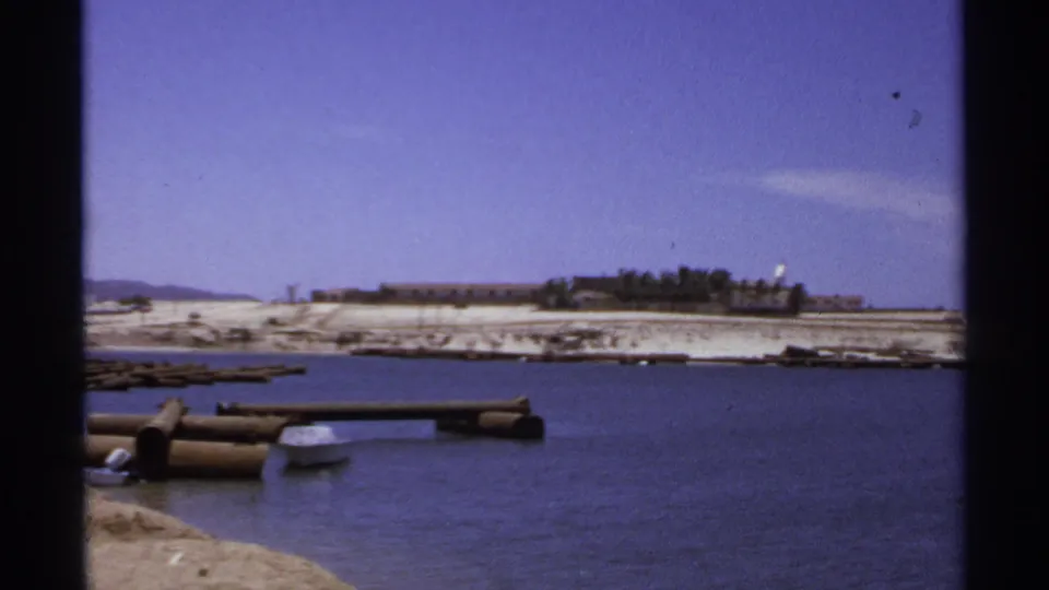 The 1974 Cabo San Lucas shoreline appears across calm water, with log docks in the foreground and low buildings in the distance.