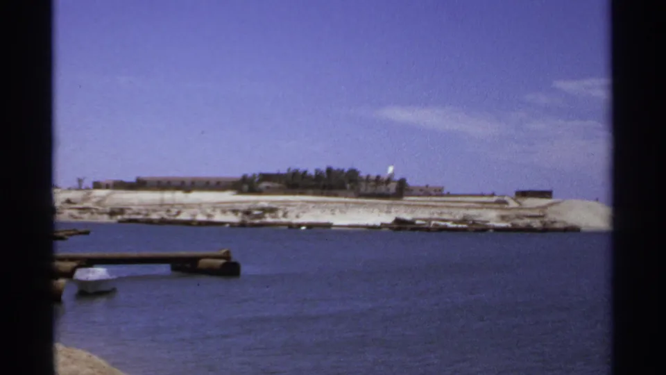 A quiet Cabo San Lucas coastal view shows dark dock posts at water level and a sandy shoreline with low buildings beyond.