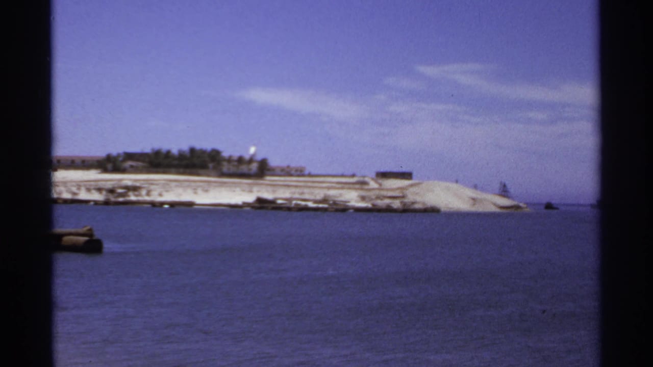 Shoreline across blue water with low buildings, pale sand, and open sky.