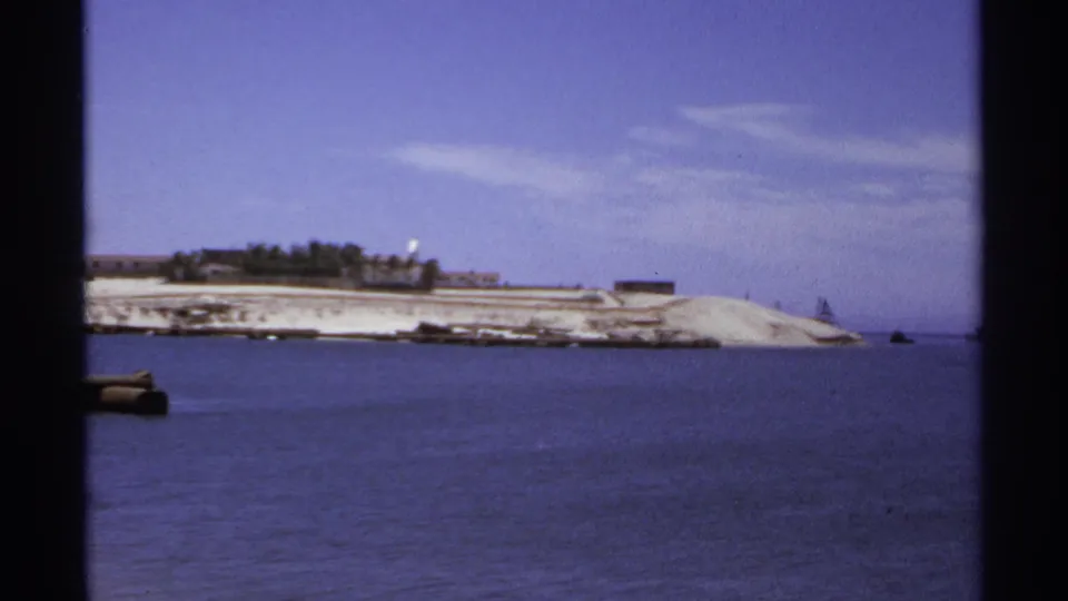 1974 Cabo San Lucas shoreline seen across calm water, with low structures, pale sand, and a clear blue sky.