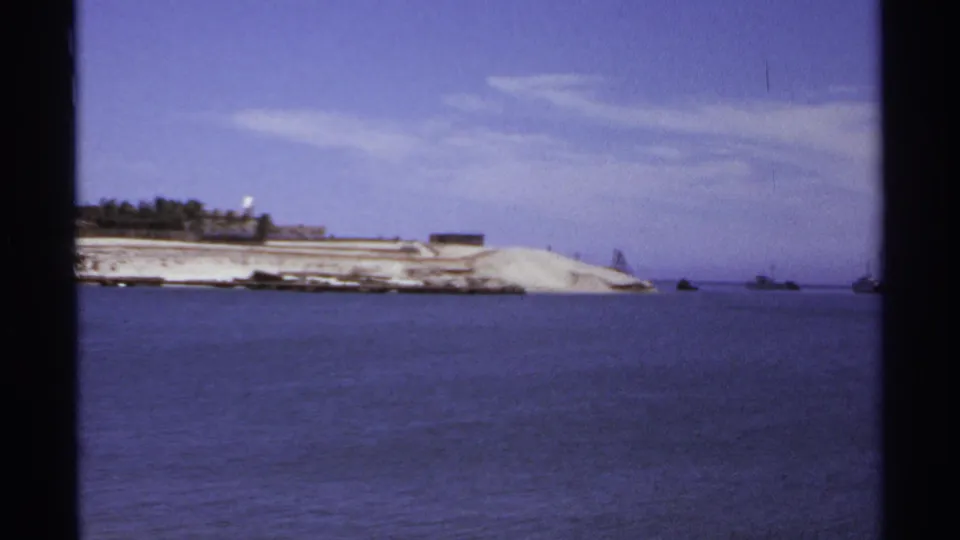 1974 Cabo San Lucas bay view with a pale shoreline, low waterfront buildings, and small distant boats offshore.