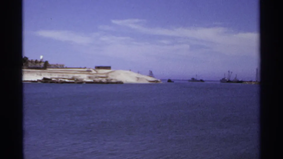 1974 Cabo San Lucas coastal frame showing calm sea, a sandy point, low shoreline buildings, and distant boats near the horizon.