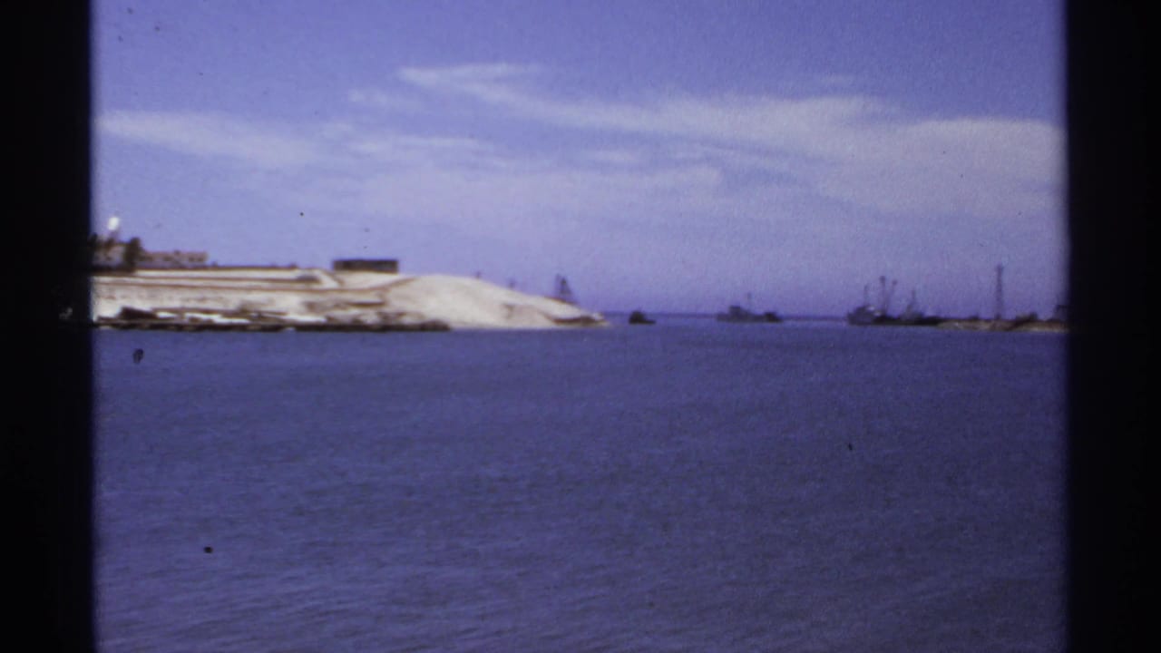 1974 Cabo San Lucas harbor view with calm water, a pale sandy shoreline, distant boats, and low waterfront structures.