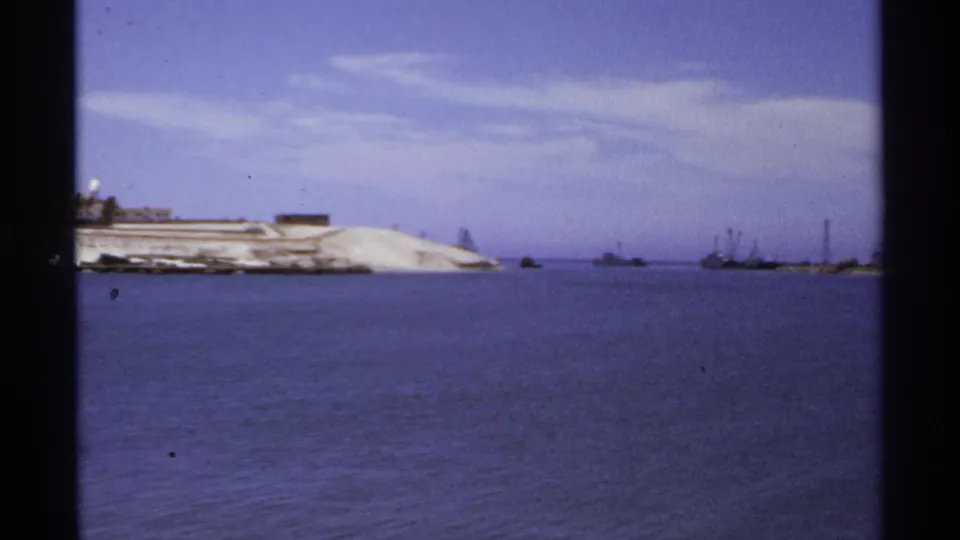 1974 Cabo San Lucas harbor view with calm water, a pale sandy shoreline, distant boats, and low waterfront structures.
