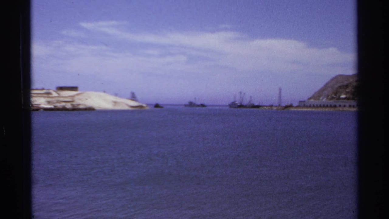 Harbor panorama with open water, sandy shore, distant vessels, and rocky hillside at right.