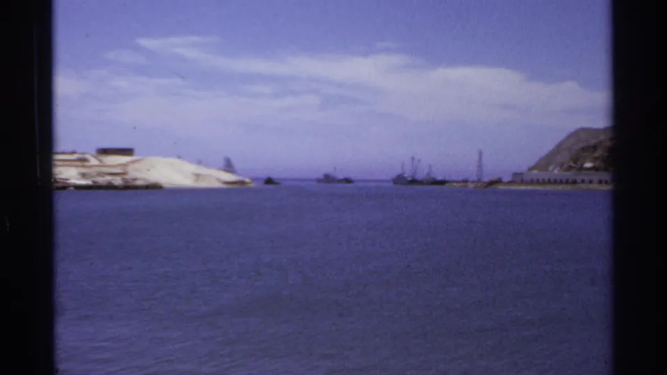 1974 Cabo San Lucas harbor panorama showing open water, distant vessels, pale shore, and a rocky hillside to the right.