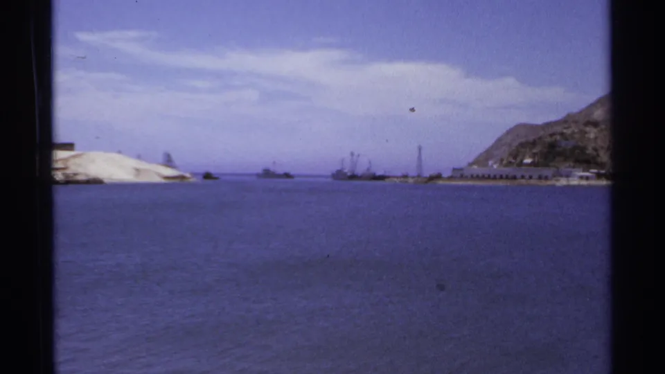 1974 Cabo San Lucas harbor view with sandy shoreline at left, distant boats near the center, and a rocky hillside at right.