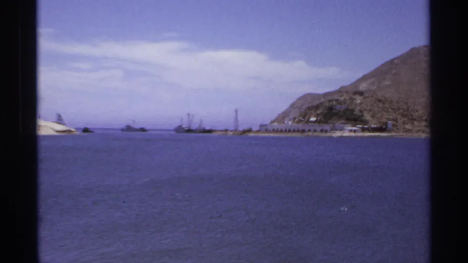 1974 Cabo San Lucas harbor scene with open blue water, small boats, low shore structures, pale sand, and a rocky hillside.