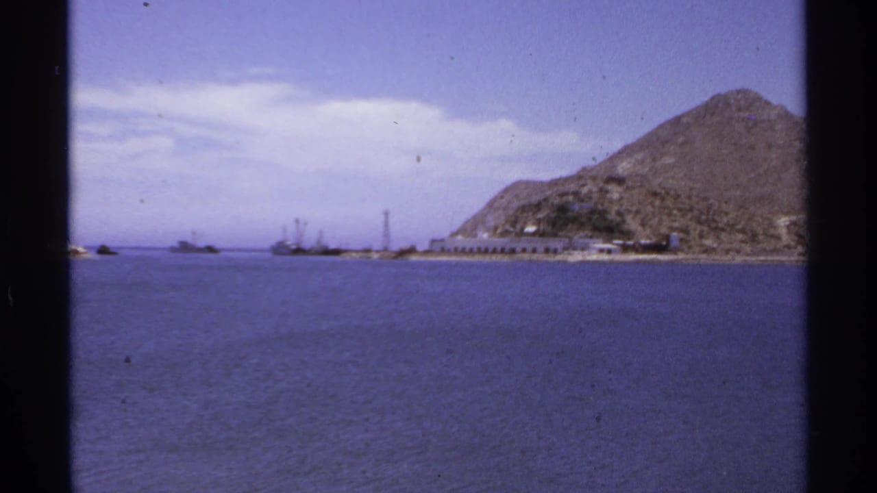 Calm bay water with rocky hills, distant boats, and a low waterfront structure.