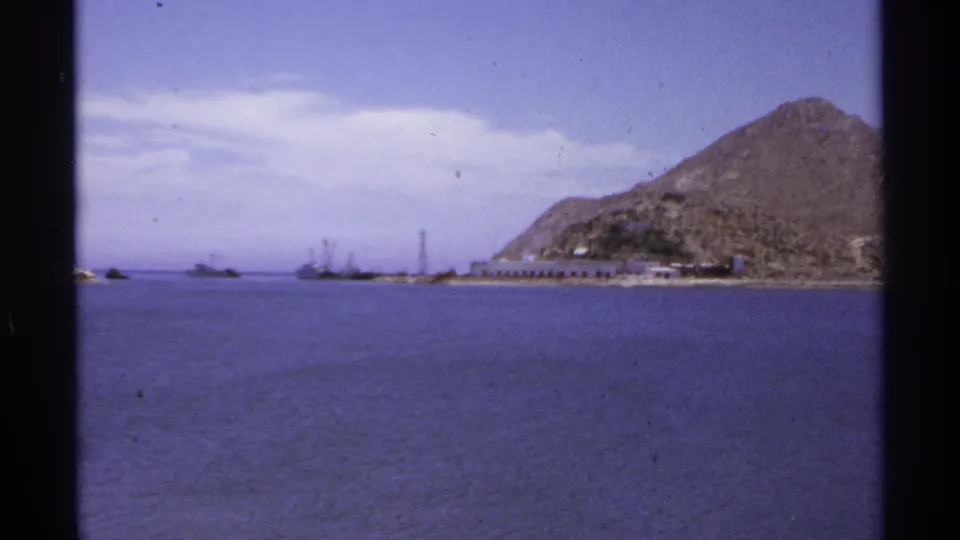 1974 Cabo San Lucas 8mm frame showing blue water, rocky hills, distant vessels, and a low pier-like shoreline structure.