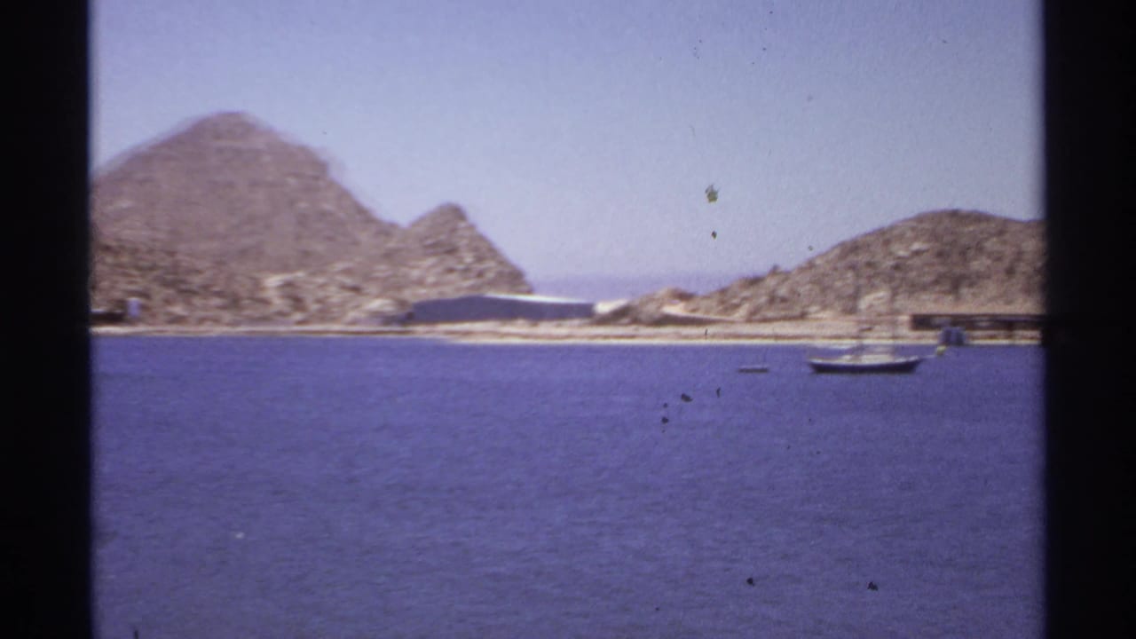 Rocky shoreline with a small sailboat on calm water and low structures near the coast.
