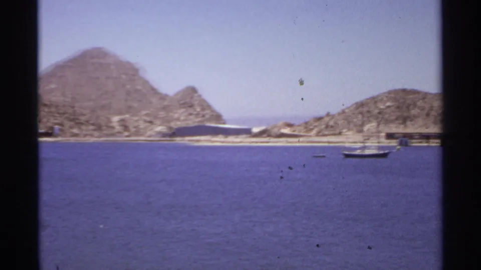 1974 Cabo San Lucas coastal view with a small sailboat near rocky shoreline, low waterfront buildings, and arid hills behind.