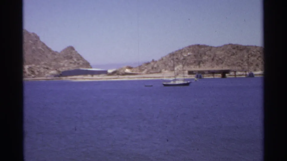 1974 Cabo San Lucas frame showing bay water, a small boat, pier structures, sandy shoreline, and rocky hills.