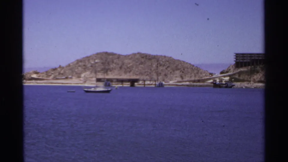 1974 Cabo San Lucas 8mm frame with a sailboat on blue water, a rocky hill, pier structures, and a waterfront building at right.