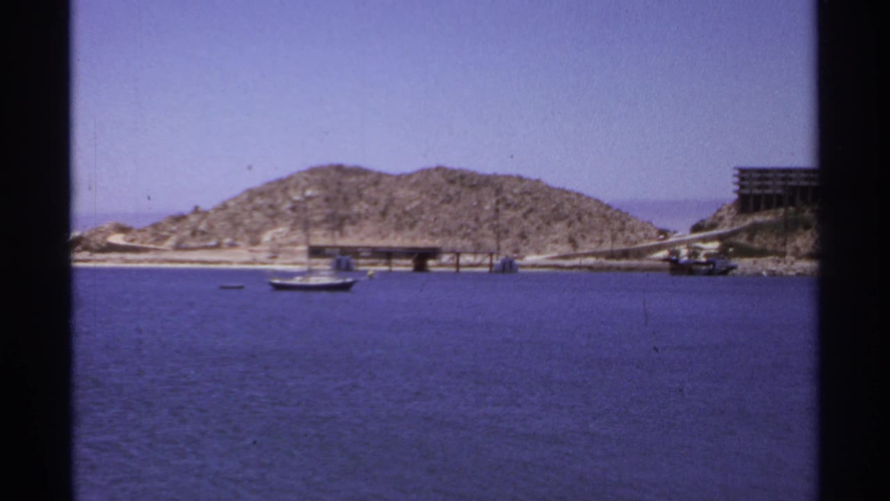 Small sailboat on blue water with rocky hill and pier in the background.