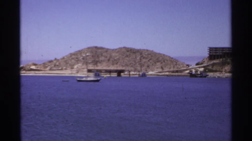 1974 Cabo San Lucas harbor view with a small sailboat, rocky hillside, wooden pier, and clear sky on 8mm film.