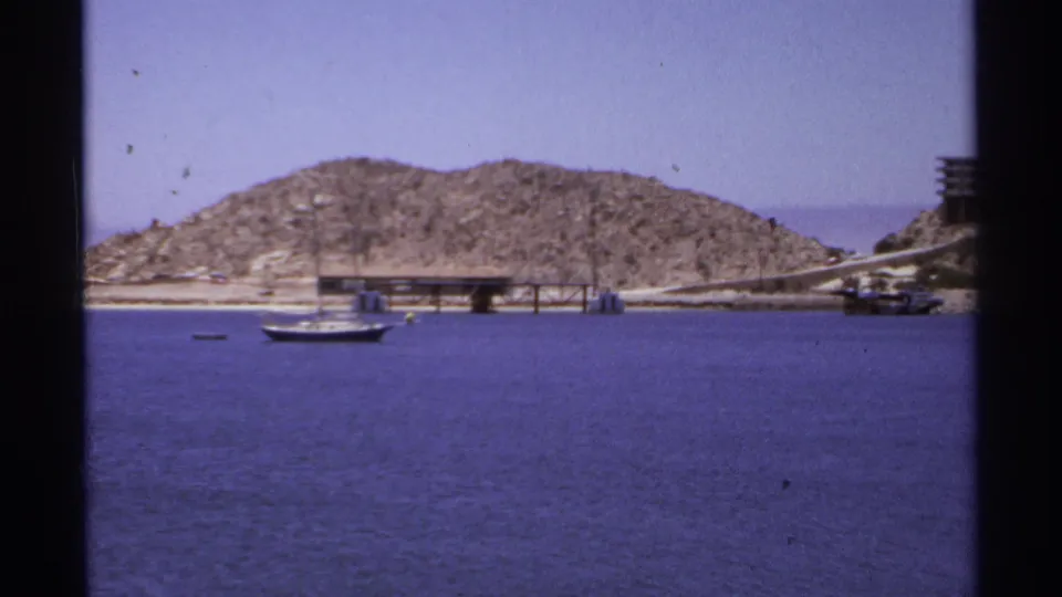 Cabo San Lucas harbor scene from 1974 showing a sailboat on blue water below a rocky shoreline and pier.
