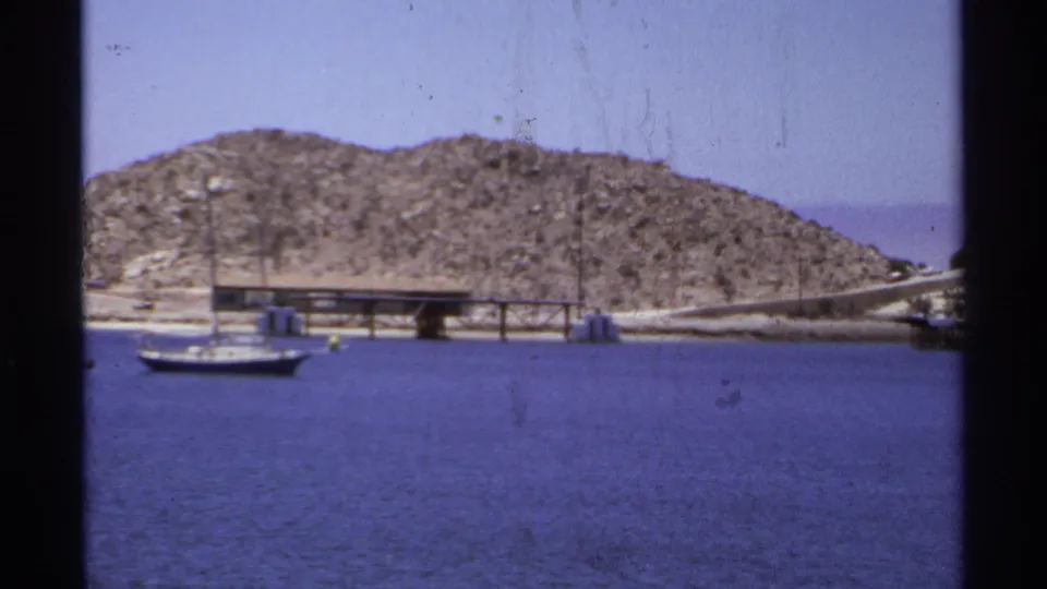Vintage 8mm frame from Cabo San Lucas in 1974 with a small sailboat, pier, rocky slope, and calm blue water.