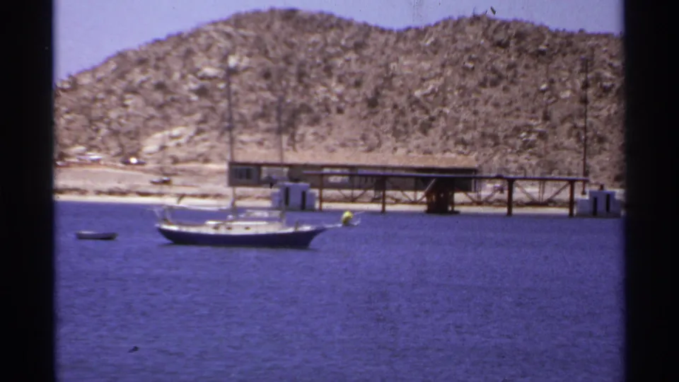 1974 Cabo San Lucas waterfront frame with a sailboat, small buoy or dinghy, wooden pier, and rocky hill beyond the water.