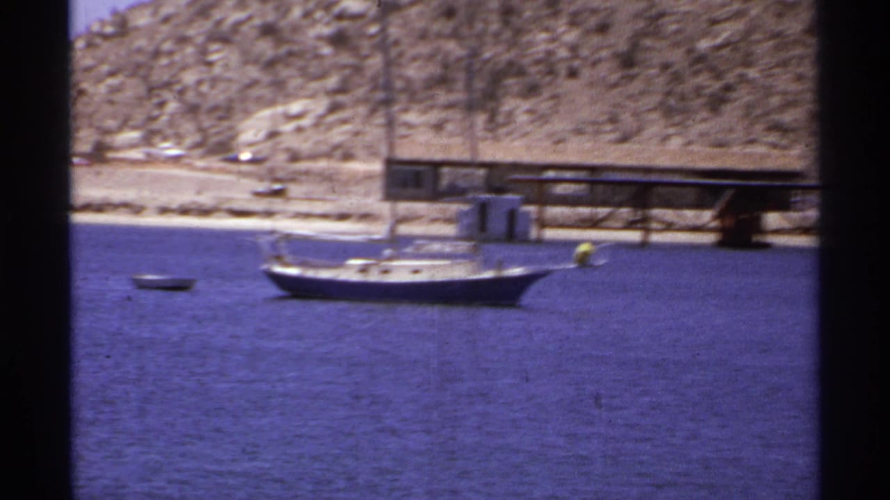 Small white sailboat anchored in blue water near a desert shoreline and pier.