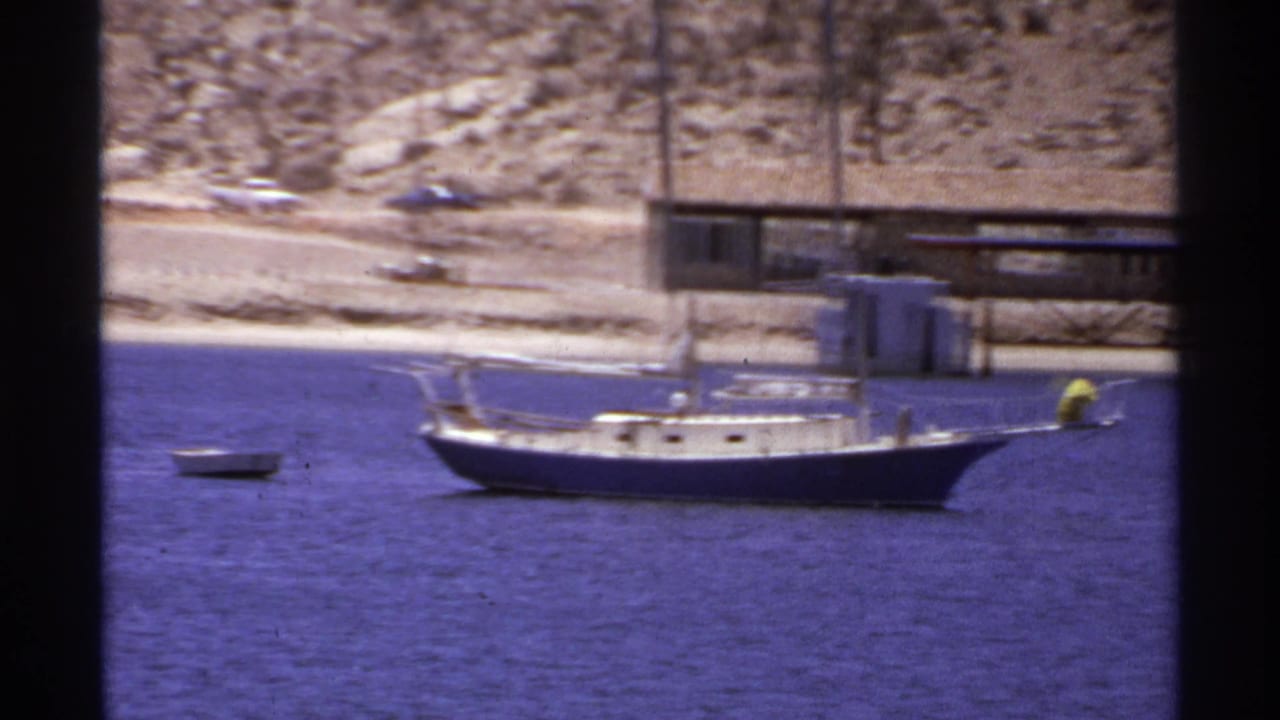 1974 Cabo San Lucas frame with the sailboat slightly closer to the pier, set against blue water and a dry rocky coastline.