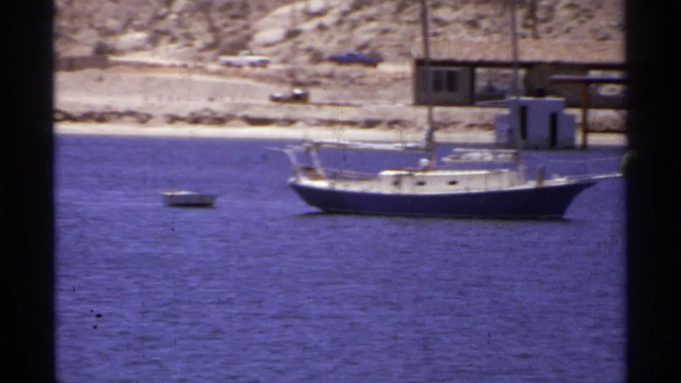 1974 Cabo San Lucas 8mm frame showing an anchored sailboat, nearby dinghy, and simple waterfront structures along a desert coast.