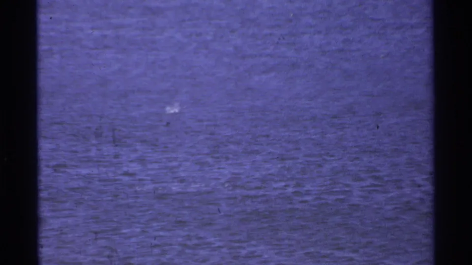 1974 Cabo San Lucas home-movie frame where the camera drifts off the sailboat, leaving mostly blue harbor water and a partial shoreline edge.