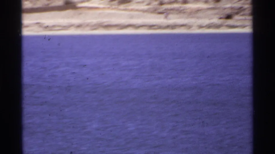 Cabo San Lucas 1974 home-movie frame with a faint sandy shoreline beyond blue-purple ocean water and visible 8mm grain.