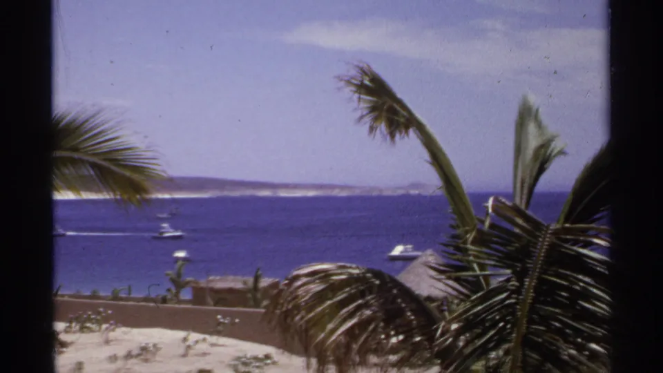 1974 Cabo San Lucas home-movie frame showing palm leaves, resort wall, small boats, blue water, and distant coastal hills.