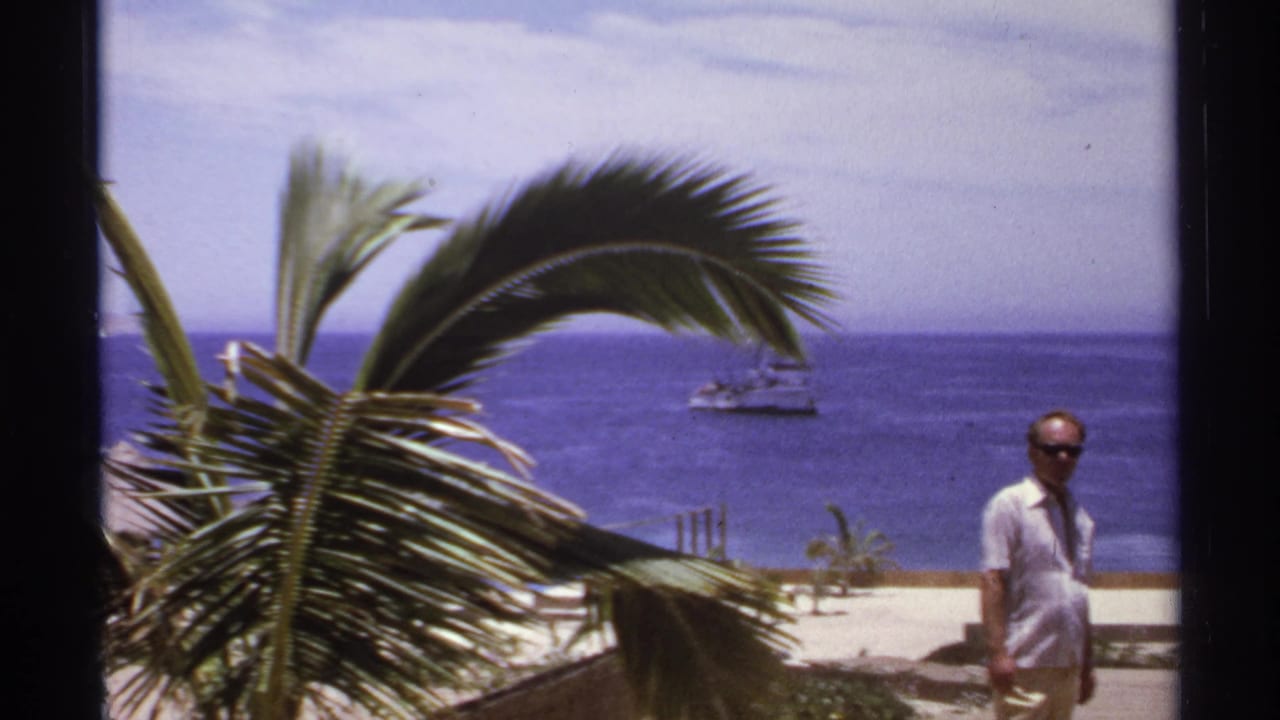 Man in sunglasses stands near a beach overlook with sea and boats behind him.