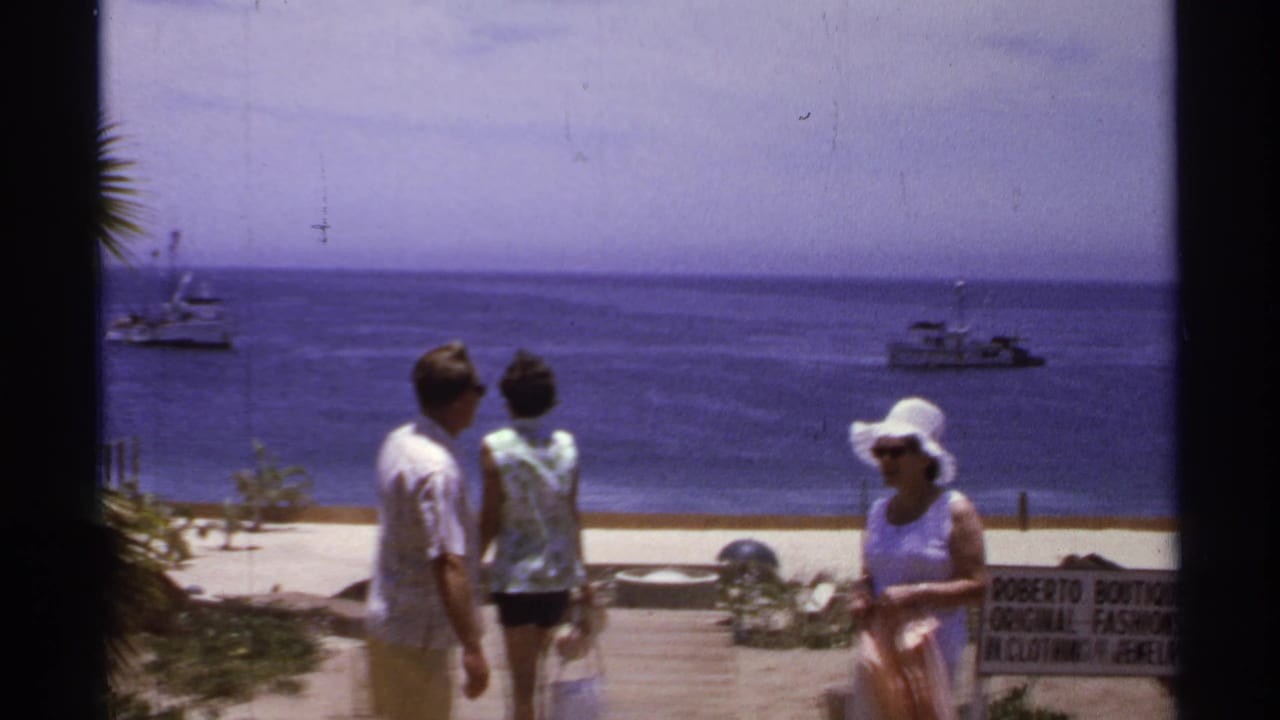 1974 Cabo San Lucas home-movie frame showing adults by the beach, blue sea, small boats offshore, and part of a Roberto Boutique sign.