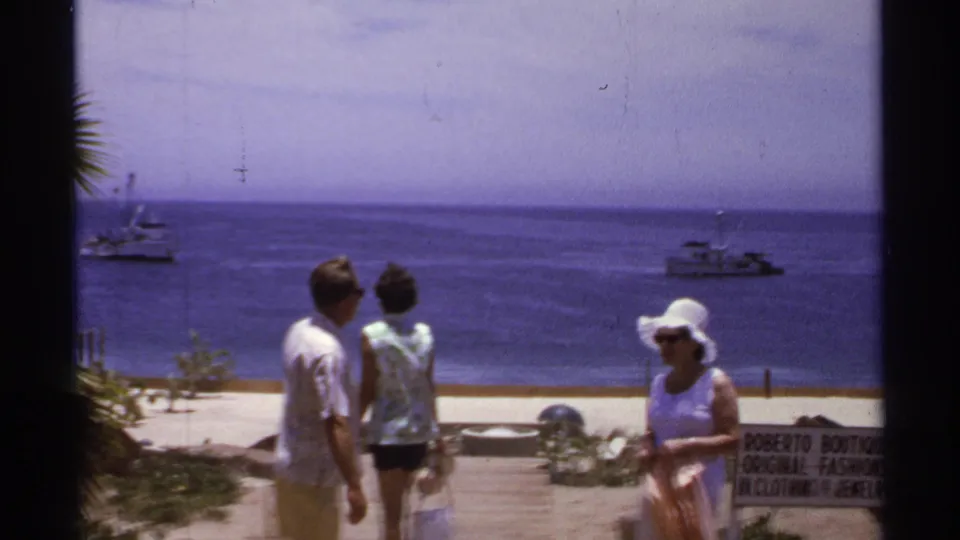 1974 Cabo San Lucas home-movie frame showing adults by the beach, blue sea, small boats offshore, and part of a Roberto Boutique sign.