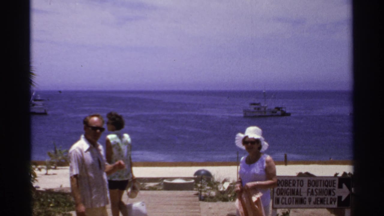 Man in sunglasses and two women near the beach with a Roberto Boutique sign and boats offshore.
